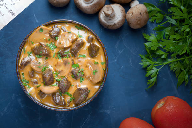 Traditional beef stroganoff with cream of champignon mushrooms in a blue bowl with its ingredients seen from above stock photo