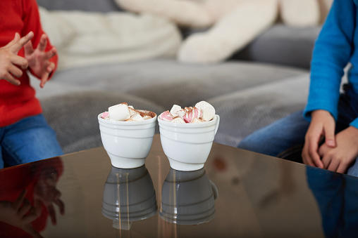 Two white cups with cocoa and marshmellows on a transparent glass table on the background of two children sitting around the table