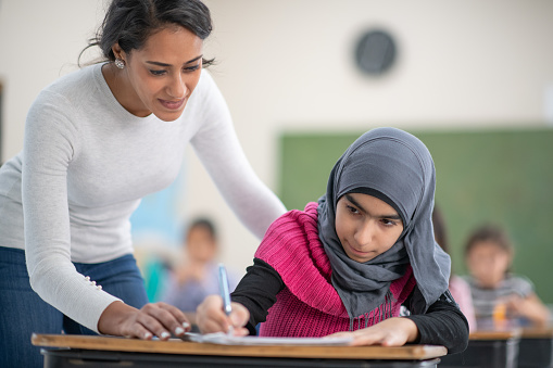 A Muslim elementary school girl wearing a hijab is writing at her desk as her teacher is next to her to check in on her work.