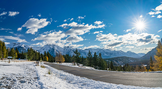 Country road in valley of forest in a snowy autumn sunny day. Colorful yellow and green trees, snow-covered mountains. Tunnel Mountain road, Banff National Park, Canadian Rockies.