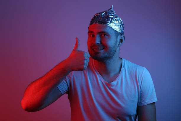 Cheerful bearded Man in foil hat smiles and throws his thumb up in red-blue neon light. Cheerful bearded Man in foil hat smiles and throws his thumb up in red-blue neon light. tin foil hat stock pictures, royalty-free photos & images