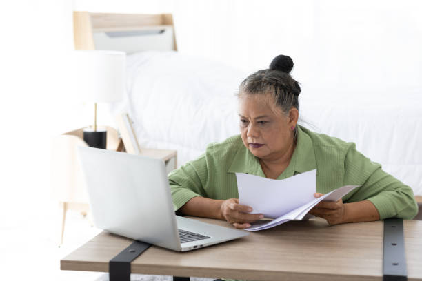 istock senior woman looking laptop computer and holding paper document at home 1296649205