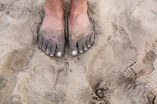 Sandy feet of a woman standing on the sand by the sea.