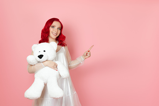 a smiling, cheerful young woman in a white dress and with red hair holds a large white teddy bear and points with her index finger at an empty space for text, isolated on a pink background.