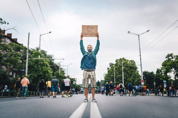 istock Young man with protective face mask, surrounded by a crowd, holding a poster in his hands. Young activist / protester against the corruption. Human rights and social issues concept. 1296673295