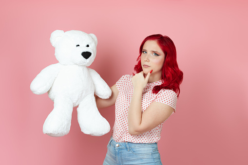 close-up brooding, suspicious young woman with red hair holds a large white teddy bear and rubs her chin with her hand , isolated on a pink background.