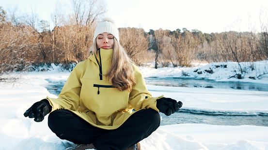 The girl sits in the lotus position in the winter in the forest and looks at the river. Meditation