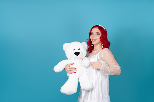 smiling happy young woman in white dress with red hair holds white teddy bear and gives thumbs up isolated on blue background.