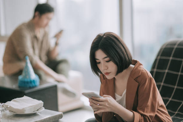 displeased asian chinese couple using phone at living room sitting apart with serious facial expression asian chinese couple using phone at living room sitting apart with serious facial expression asian couple trouble stock pictures, royalty-free photos & images