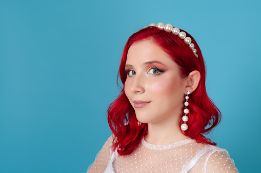 Close-up face of a young woman with red wavy hair in a white dress, pearl headband and long dangling earrings, isolated on a blue background.