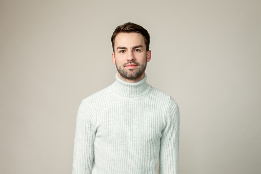 Studio portrait of attractive 20 year old bearded man in white sweater on gray background