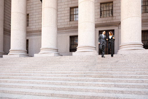 A lawyer  and their client  or business people walk down the staircase of a courthouse or municpal building. They are smiling and chatting.