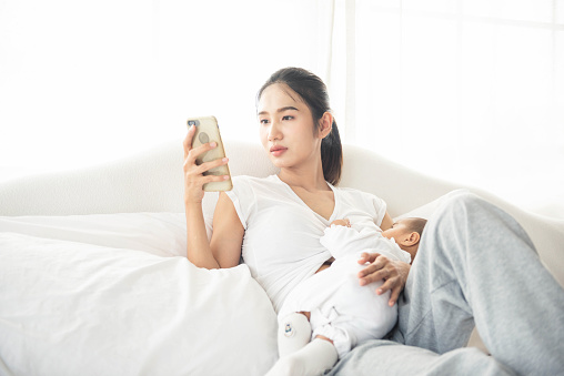 Young asian mother feeding breast her baby on bed at home in white room. Asia mom holding her baby child and use smartphone to check email. Woman and new born relax and use cellphone in bedroom.
