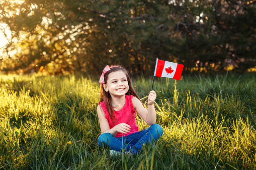 Adorable cute happy Caucasian girl holding Canadian flag. Smiling child sitting on grass in park holding Canada flag. Kid citizen celebrating Canada Day holiday on first day of July outdoor.