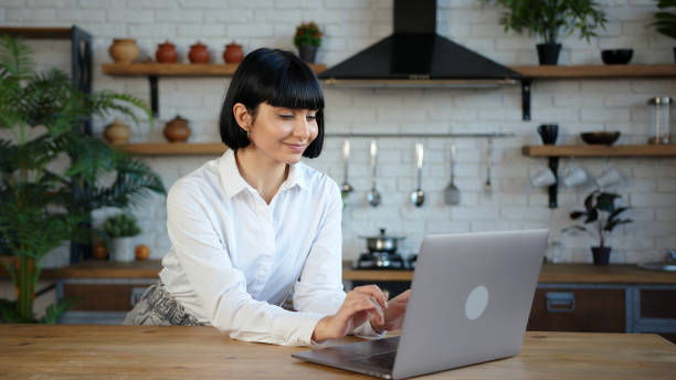 istock Lady in white blouse works on laptop at table in kitchen 1296715083