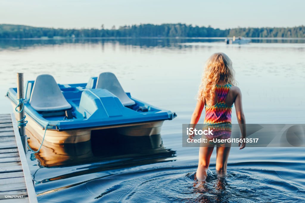 Cute blonde girl walking to pedal boat in lake water on sunset. Summer sport water outdoors activity. Real authentic happy childhood lifestyle. View from back behind. Cute blonde girl walking to pedal boat in lake water on sunset. Summer sport water outdoor activity. Real authentic happy childhood lifestyle. View from back behind. Child Stock Photo