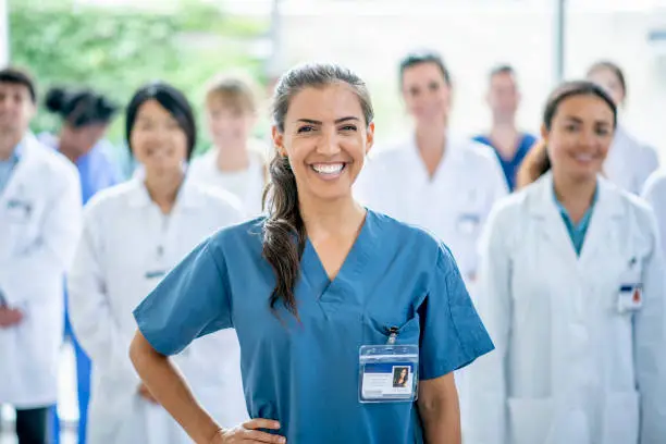 A female medical student of Hispanic ethnicity smiles for the camera as she stands in front of her group of medical professionals in the background.
