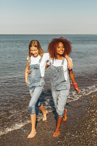 Two young women holding hands walking by the beach on their summer vacation and they smile and enjoy their vacation.