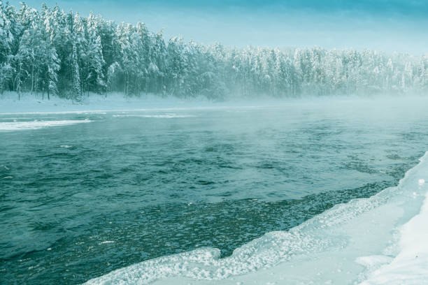 Frosty fog over winter river with snow and forest on bank. First ice on lake on cold day. stock photo