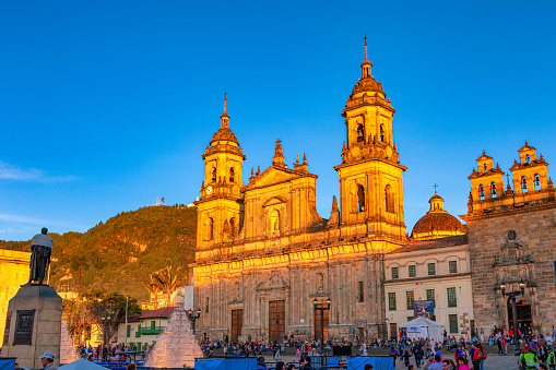 Looking from Plaza Bolivar, in the Andean Capital city of Bogota, in Colombia, South America, to the North Eastern corner of the Square. The 19th Century Catedral Primada has the upper half turned to gold by the setting sun. The Cathedral is the seat of the Roman Catholic Archbishop of Colombia. A few metres to the right, is the Archbishop's chapel. A section of his palace can also be seen. The style of architecture is classical colonial Spanish. In the far background is the mountain peak of Monserrate. The altitude at street level is about 8,500 feet above mean sea level. Image shot in the late afternoon sunlight; horizontal format. Copy space.