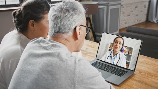 Back view of aged couple looking at screen of laptop and talking to doctor woman using online video call, medical consultation at home
