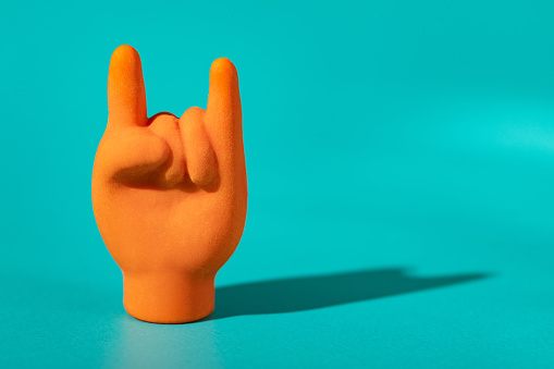 orange sign of the horns hand gesture, rock & roll hand sign icon on bright teal background with hard shadows