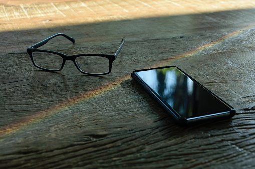 Glasses and smartphone on a rustic wooden table. Sunbeam causing a scattering of light, forming a rainbow.