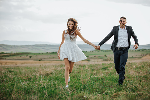 Cheerful young couple holding hands, smiling and happily running on meadow, enjoying love and freedom in nature, front view