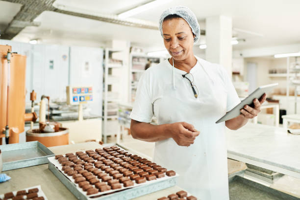 Worker using a digital tablet in a commercial chocolate making factory Smiling chocolate factory worker using a digital tablet while checking a tray of freshly made confectioneries baker occupation stock pictures, royalty-free photos & images
