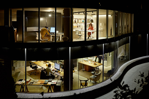 Bi-level exterior view of design professionals photographed through building windows as they work independently at computers in modern office.