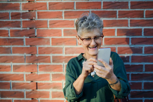 istock Senior woman standing outdoors against brick wall background, using smartphone. 1296814144