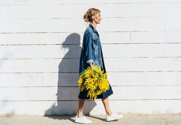 Stylish young woman in denim clothes with a bouquet of mimosa Stylish young woman in denim clothes with a bouquet of mimosa walks along the wall sanctified by the bright spring sun. march month photos stock pictures, royalty-free photos & images