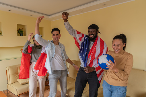 A Group of Friends of Different Ethnicities from the USA and Canada is Sitting at Home Enjoying in Sports Game on Television Program. Cheerful Friends Support Soccer Teams and Using National Flags.