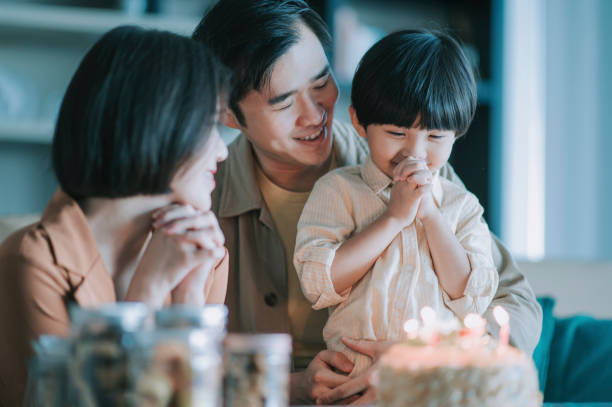 asiatische chinesische familie augen geschlossen machen geburtstagswünsche vor geburtstagstorte vor dem blasen geburtstagskerze im wohnzimmer feiern sohn geburtstag - kinderwunsch stock-fotos und bilder