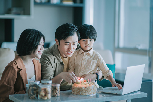 asian chinese family lighting birthday candle getting ready to videocall grandparent with birthday celebration in living room