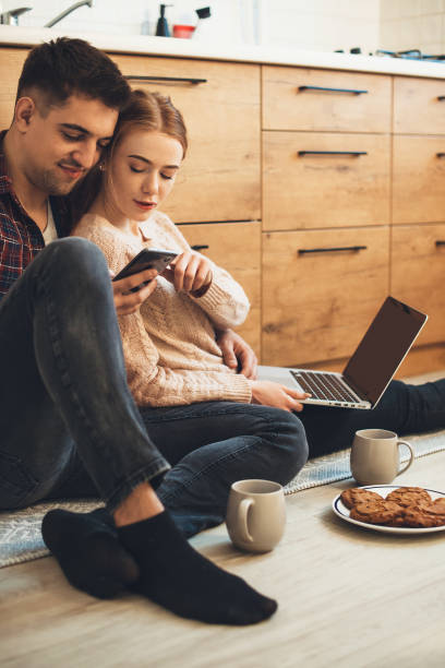 istock Cheerful couple sitting on the floor in the kitchen using a laptop and phone while drinking tea with cakes in the morning 1296835454