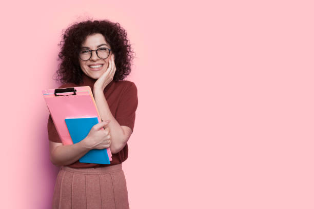 istock Surprised curly haired student is touching her cheek smiling on a pink wall with free space holding some folders 1296838037
