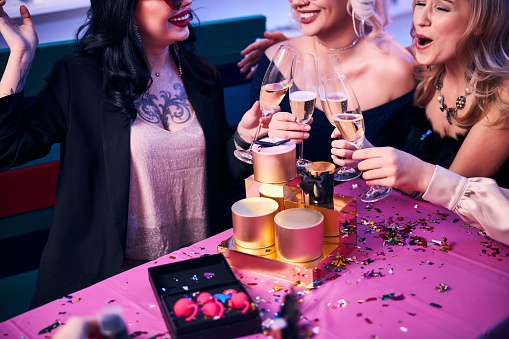 Cropped photo of smiling joyous female friends clinking their champagne flutes at the festive table