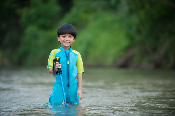 Asian kids inside the river smiling at the camera. His hand holding one stick. He wear a turquoise swimming suit.