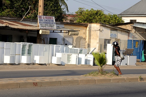 Avepozo, Togo - November 30, 2019: Woman wearing a motorbike helmet stands at the highway. Location: Avepozo, Togo, West Africa. A shop is selling used fridges and freezers.