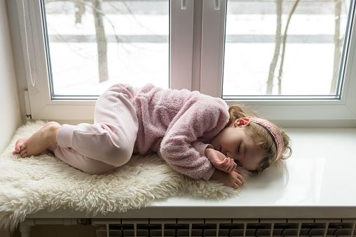 Kid lies on fur rug on windowsill at home, little girl dreams inside house in winter. Child sleeps on room sill in white interior. Concept of quarantine due to COVID-19, childhood safety and sleeping.