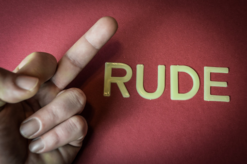 Human middle finger beside the word Rude written with plastic letters on red paper background, offense sign concept