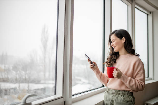 istock Woman drinking coffee, using her phone and day dreaming while looking through window 1296876923