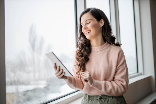 istock Beautiful young woman using her digital tablet and standing by window at home 1296877007