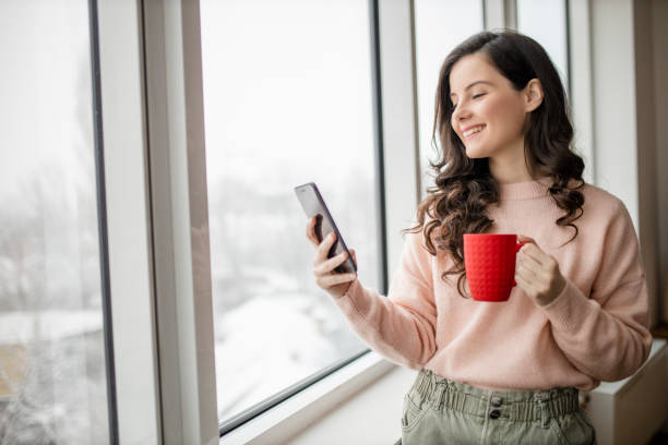 istock Woman drinking coffee, using her phone and day dreaming while looking through window 1296877282
