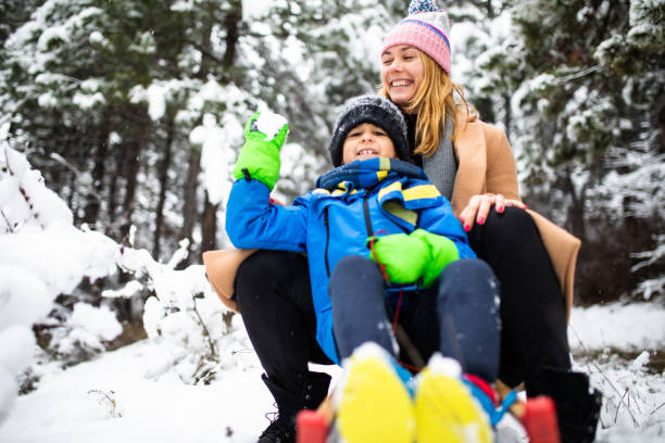 istock A cute boy about to throw a snowball at the camera while sitting on a sled with his mom 1296878402