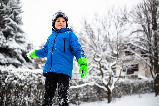 A cute boy with his clothes covered in snow is about to throw a snowball out in his yard on a snowy day