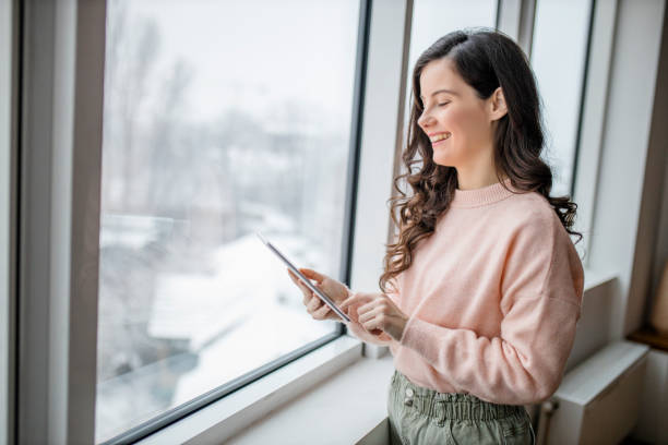istock Beautiful young woman using her digital tablet and standing by window at home 1296879882