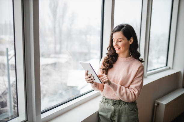 istock Beautiful young woman using her digital tablet and standing by window at home 1296881650