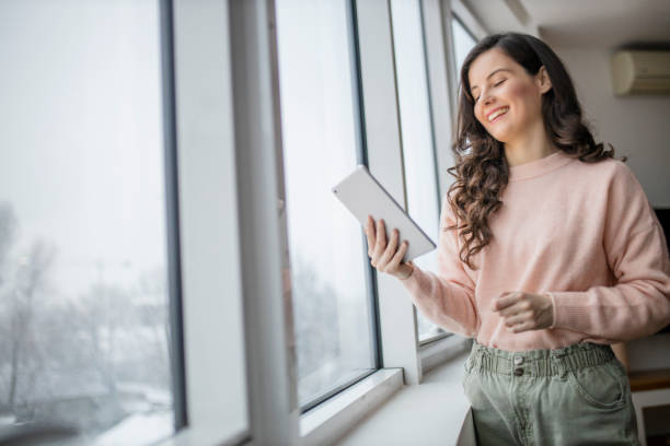 istock Beautiful young woman using her digital tablet and standing by window at home 1296882123